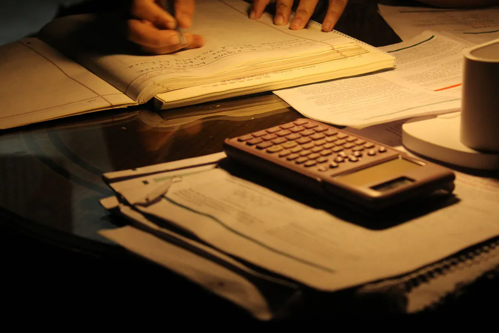 A calculator and paper records on a desk under a lamp at night while someone catches up on the books