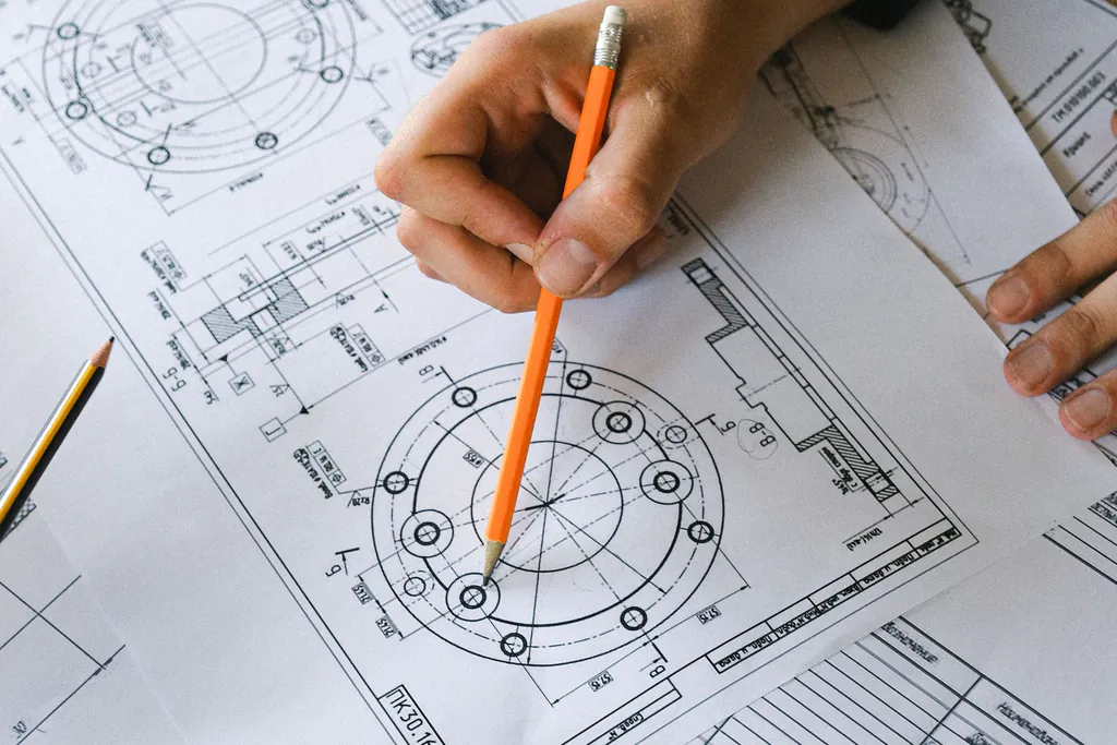 A pencil traces a hole pattern on a printed part drawing, checking each feature one by one