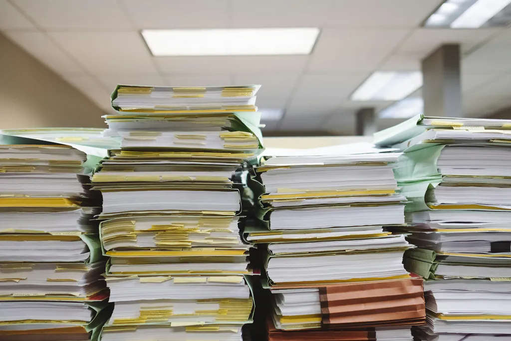 Tall stacks of paper files and folders piled on a desk, where a new request could easily get buried