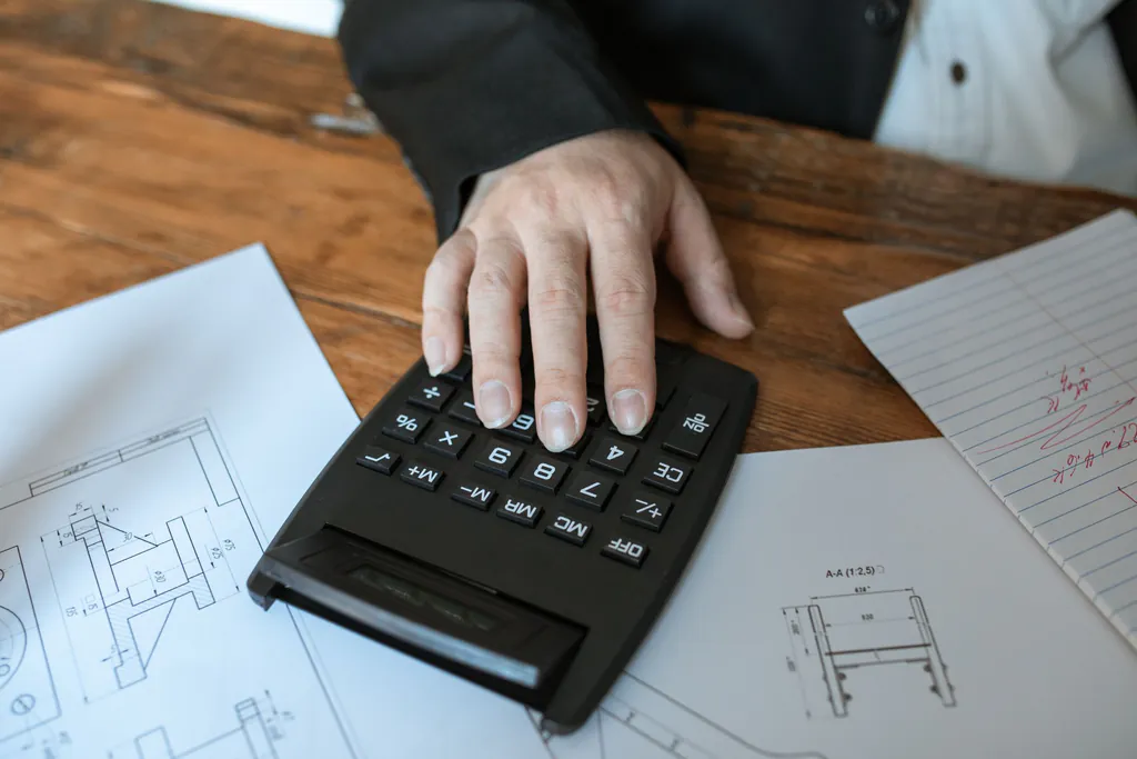 A hand on a calculator over printed part drawings, working out the price for a quote