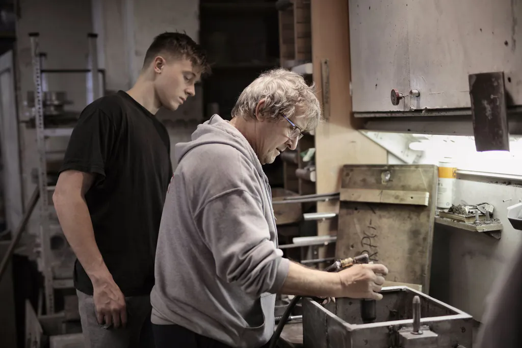 An older machinist shows a young apprentice how he works a part at the bench