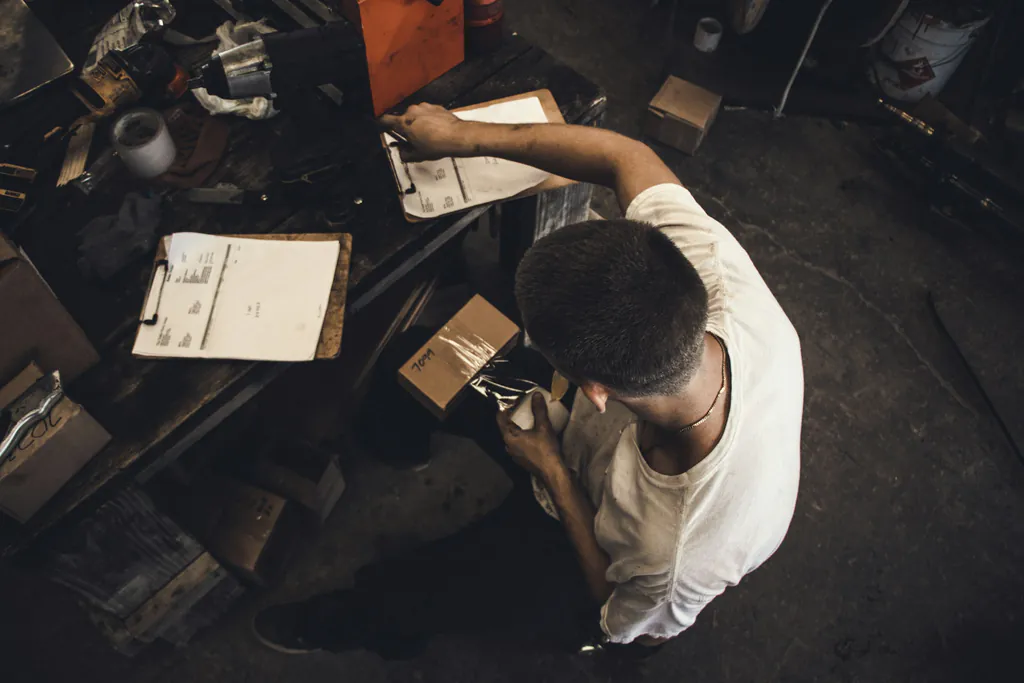 A shop worker packs a boxed part beside clipboards of order paperwork on a cluttered workbench
